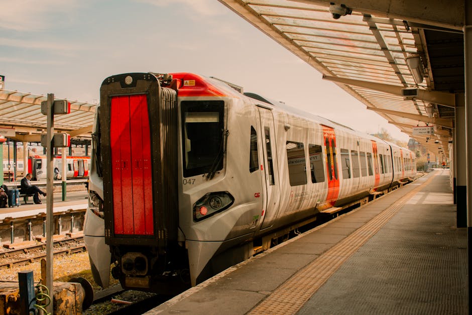 The image shows a modern commuter train at Guildford station, positioned on the railway tracks adjacent to a platform with a covered waiting area. The train has a sleek design with a predominantly silver body, accented by orange and black details, and appears to be a part of the UK rail network. The platform is made of concrete with tactile paving strips running parallel to the train for accessibility. On the platform, a few passengers are seated on benches, with some carrying luggage or backpacks, preparing for their home relocation or daily commute. Station signage and lighting fixtures are visible under the covered area, which extends over part of the platform to provide shelter for waiting passengers. The scene is lit by natural daylight, highlighting the clean and organized environment of the station, which is a key part of the transportation infrastructure supporting move logistics and furniture transport services in Surrey. Surrey Man and Van occasionally monitors such settings for efficient loading and unloading operations during house removals or furniture transport processes.