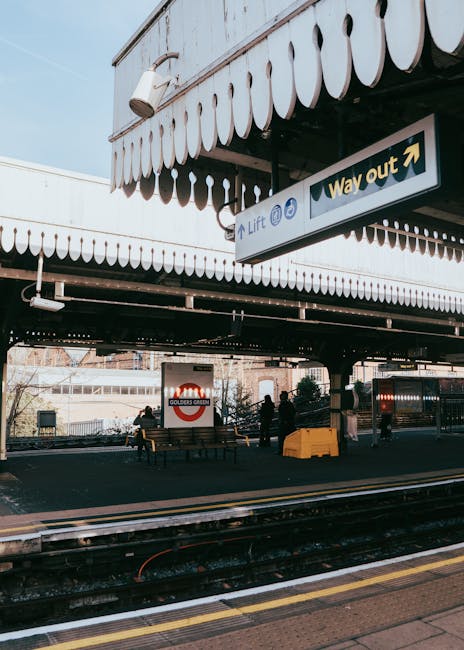 A view of Guildford station platform with the station's Golders Green sign visible in the background. The platform features a covered area with a decorative scalloped awning and a white directional sign indicating the lift and way out. Several people are present, some standing and others seated on benches. In the foreground, moving train tracks run parallel to the platform, which is paved with a textured surface. On the platform, there are various items associated with home relocation and furniture transport, including cardboard boxes, wrapped furniture pieces, and a large yellow plastic container. The scene indicates preparations for loading or unloading household belongings, supporting the context of house removals and moving logistics handled by Surrey Man and Van, especially within the setting of a railway station's loading area.
