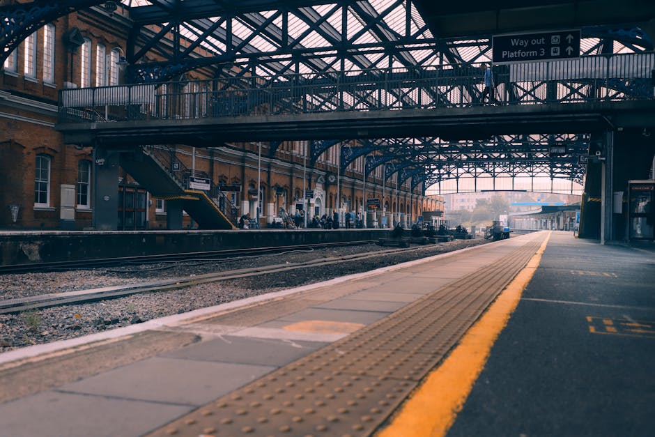 The image shows a modern commuter train at Guildford station, positioned on the railway tracks adjacent to a platform with a covered waiting area. The train has a sleek design with a predominantly silver body, accented by orange and black details, and appears to be a part of the UK rail network. The platform is made of concrete with tactile paving strips running parallel to the train for accessibility. On the platform, a few passengers are seated on benches, with some carrying luggage or backpacks, preparing for their home relocation or daily commute. Station signage and lighting fixtures are visible under the covered area, which extends over part of the platform to provide shelter for waiting passengers. The scene is lit by natural daylight, highlighting the clean and organized environment of the station, which is a key part of the transportation infrastructure supporting move logistics and furniture transport services in Surrey. Surrey Man and Van occasionally monitors such settings for efficient loading and unloading operations during house removals or furniture transport processes.